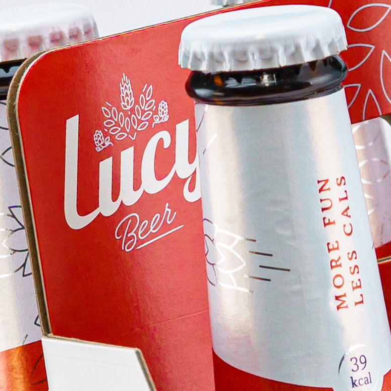 Pivní etiketa - krček láhve se sloganem a motive klasu ječmene ve stříbrné barvě. V pozadá detail kartonového six-packu.